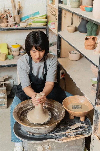 Joven mujer asiática artesana en delantal moldeado arcilla mojada en la rueda de cerámica cerca de cuenco con agua y herramientas mientras trabaja en taller de cerámica, herramientas de cerámica y equipo - foto de stock