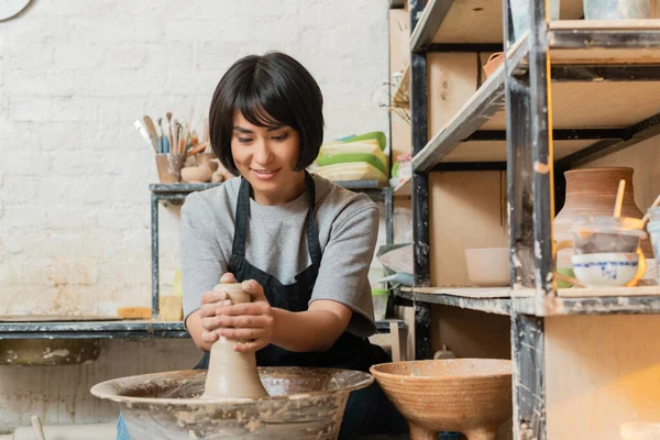 Sonriente joven artista femenina asiática en delantal moldeado de arcilla en la rueda de cerámica mientras trabaja cerca de rack y esculturas en taller de cerámica en el fondo, herramientas de cerámica y equipo - foto de stock