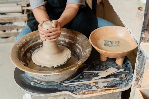 Cropped view of young female artisan in apron molding wet clay on pottery wheel near bowl with water, sponge and tools on table in ceramic workshop, pottery tools and equipment — Stock Photo