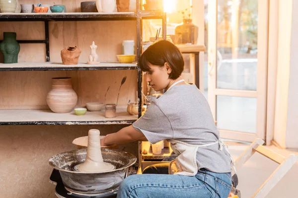 Side view of young brunette asian artisan in apron working with wet clay on pottery wheel and bowl with water in ceramic workshop at sunset, pottery tools and equipment — Stock Photo