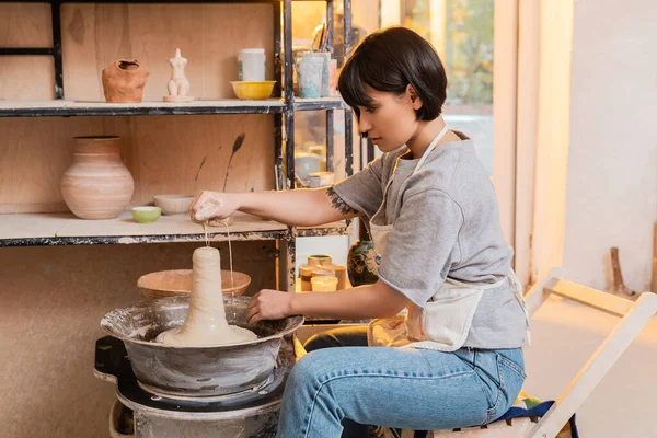 Side view of young asian female artisan in apron pouring water from sponge on wet clay on pottery wheel and working in ceramic workshop at sunset, pottery tools and equipment — Stock Photo