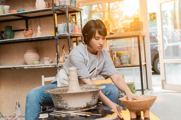 Young asian brunette artist in apron taking bowl with water near clay on pottery wheel and tools in ceramic workshop at background at sunset, pottery tools and equipment — Stock Photo