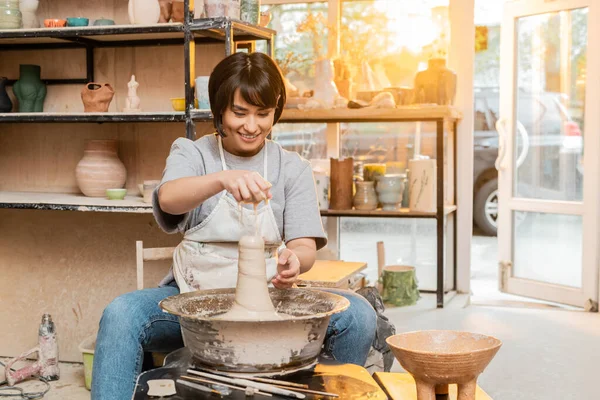 Jeune artiste asiatique souriante dans un tablier versant de l'eau sur de l'argile sur une roue de poterie près d'outils et un bol dans un atelier flou au coucher du soleil, un artisan créant des pièces de poterie uniques — Stock Photo