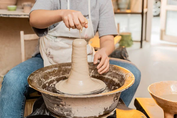 Обрізаний вид жіночої майстрині в фартусі, що поливає воду на глину, працюючи на гончарному колесі в розмитій керамічній майстерні на фоні, майстер створює унікальні керамічні деталі — стокове фото
