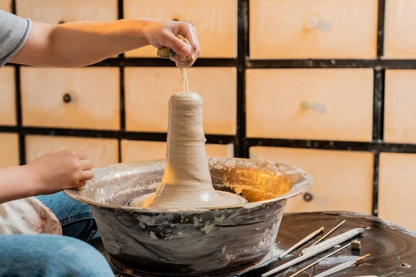 Vista cortada de oleiro fêmea jovem derramando água de esponja em barro enquanto trabalhava na roda de cerâmica perto de ferramentas em oficina no fundo, artesão criando peças de cerâmica únicas — Fotografia de Stock