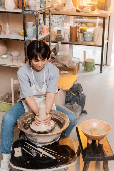 Young brunette asian artisan in apron molding wet clay on pottery wheel near bowl with water and tools in blurred ceramic workshop at sunset, artisan creating unique pottery pieces — Stock Photo