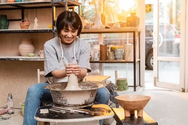 Весела молода азіатська жінка-гончарка в фартухах, що формує глину на гончарному колесі біля миски та інструментів в керамічній студії на фоні заходу сонця, створює унікальні керамічні деталі — стокове фото