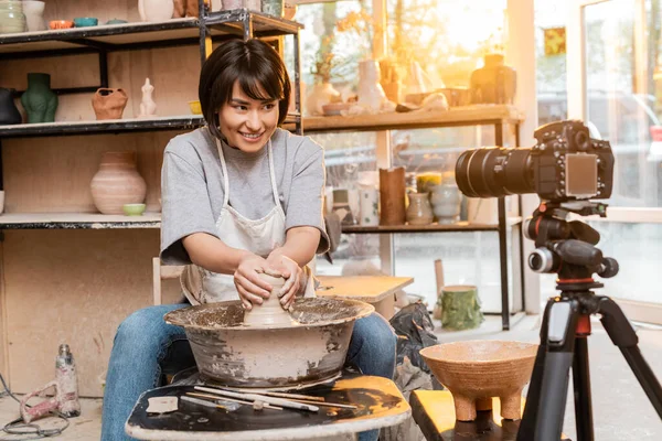 Young and cheerful asian artist in apron molding clay on pottery wheel near tools and looking at blurred digital camera on tripod in workshop at sunset, artisan creating unique pottery pieces — Stock Photo