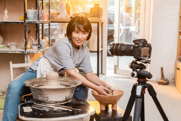 Positive young asian female artist in apron talking to digital camera on tripod and holding sponge near bowl with water and clay on pottery wheel in studio at sunset, artisan creating pottery pieces — Stock Photo