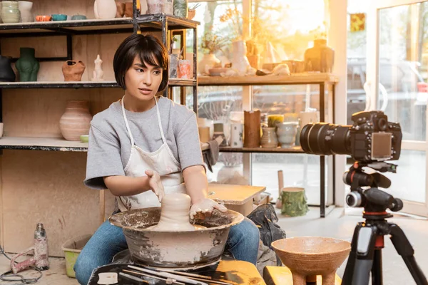 Jeune artisan asiatique en tablier façonnant de l'argile humide sur roue de poterie et parlant à un appareil photo numérique sur trépied dans un atelier de céramique floue en arrière-plan, artisan créant des pièces de poterie uniques — Photo de stock