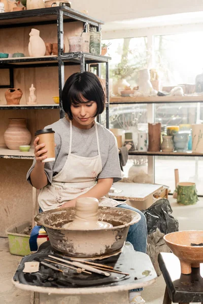 Jeune potier asiatique dans un tablier tenant du café à emporter et regardant de l'argile sur une roue de poterie près d'outils et d'un bol dans un atelier de poterie floue, un artisan créant des pièces de poterie uniques — Photo de stock