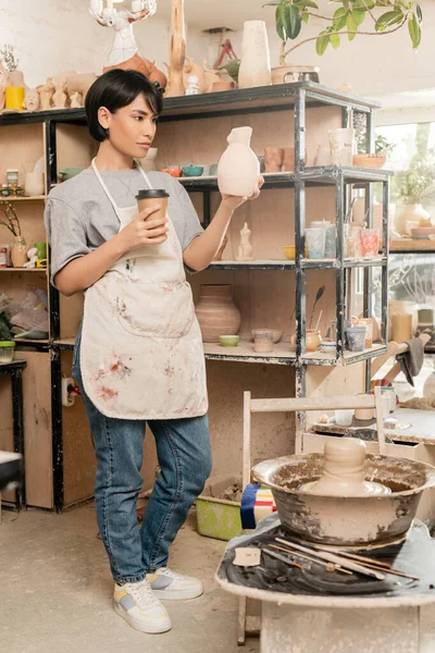 Jovem asiática artista feminina em avental segurando escultura e café para ir perto de barro na roda de cerâmica, ferramentas e rack em oficina de cerâmica, artesão criando peças de cerâmica únicas — Fotografia de Stock