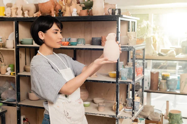 Young brunette asian female artisan in apron holding clay sculpture near rack with shelves in blurred ceramic workshop at background at sunset, artisan creating unique pottery pieces — Stock Photo