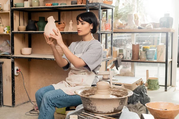 Молодая азиатская художница в фартуке, держащая керамическую скульптуру, сидя рядом с глиной на гончарном колесе и инструментах в размытой студии на закате, концепция процесса скульптуры из глины — стоковое фото