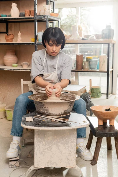 Young asian female potter in apron molding wet clay on pottery wheel near tools and bowl with sponge while working in ceramic studio at background, clay sculpting process concept — Stock Photo