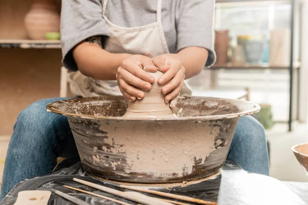 Cropped view of tattooed female artisan in apron molding wet clay and working with pottery wheel near blurred tools on table in blurred ceramic workshop, clay sculpting process concept — Stock Photo