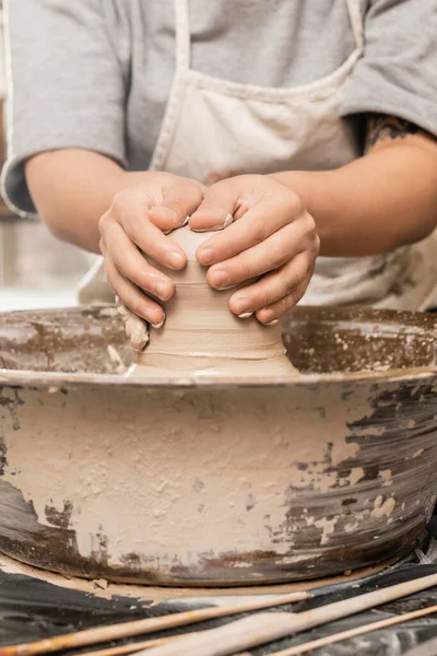 Ausgeschnittene Ansicht einer verschwommenen und tätowierten Kunsthandwerkerin in Schürze, die nassen Ton gießt, während sie an der Töpferscheibe in der Nähe von Werkzeugen auf dem Tisch im Keramik-Atelier arbeitet, Tonbildhauerei-Konzept — Stockfoto