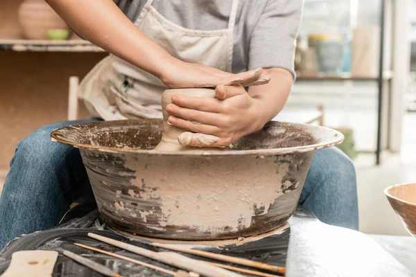Cropped view of blurred female artist in apron shaping wet clay and working on pottery wheel near wooden tools on table in ceramic workshop, clay sculpting process concept — Stock Photo