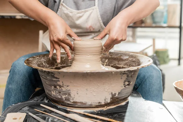 Обрізаний вид розмитих жінок-художниць у фартусі, що формує мокру глину на керамічному колесі біля інструментів на столі в керамічній художній майстерні на фоні, концепція процесу скульптури глини — стокове фото