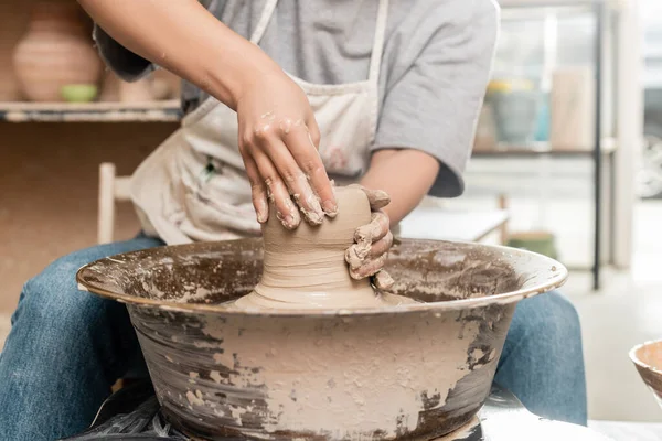 Ausgeschnittene Ansicht einer verschwommenen jungen Kunsthandwerkerin in Schürze, die Ton auf Töpferscheibe gießt, während sie im Keramik-Atelier im Hintergrund arbeitet, Konzept des Tonbildhauerprozesses — Stockfoto