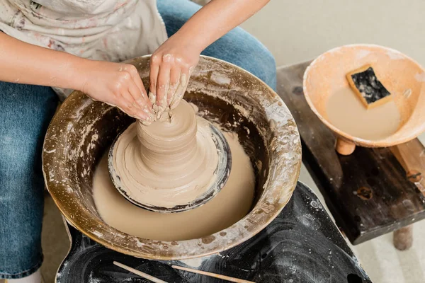 Vue grand angle de jeune artisan femelle en tablier moulant de l'argile humide sur une roue de poterie et travaillant près d'un bol flou avec de l'eau et une éponge dans un atelier de céramique, concept de fabrication de poterie habile — Photo de stock
