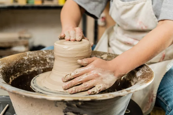 Ausgeschnittene Ansicht einer verschwommenen Kunsthandwerkerin in Schürze, die nassen Ton formt und im Hintergrund mit der Töpferscheibe in der Keramikwerkstatt arbeitet, geschicktes Töpferkonzept — Stockfoto