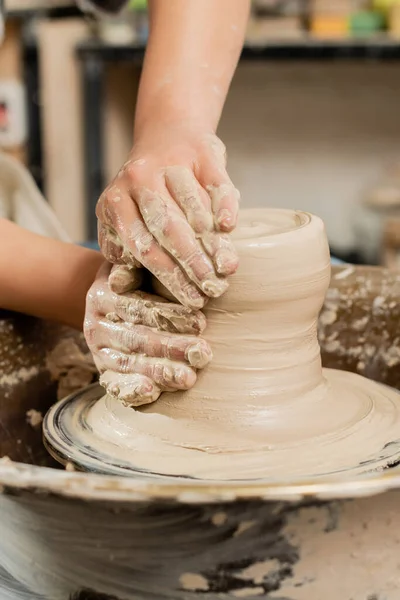 Vue recadrée du jeune céramiste femelle moulant de l'argile humide sur une roue de poterie en rotation tout en travaillant dans un atelier de céramique floue en arrière-plan, concept de fabrication de poterie qualifiée — Photo de stock