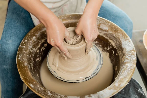Vue grand angle du jeune potier femelle en tablier façonnant de l'argile humide tout en travaillant sur la rotation de la roue de poterie dans un atelier d'art en arrière-plan, concept de fabrication de poterie qualifiée — Photo de stock