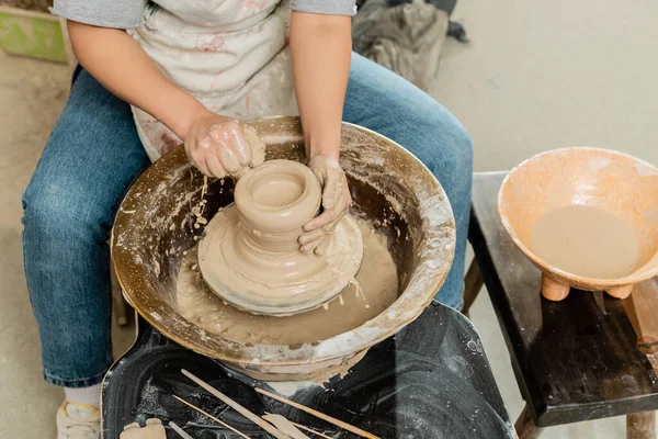 Высокий угол обзора молодой женщины-гончара в фартуке, льющейся водой на глину и крутящееся гончарное колесо возле чаши и инструменты в художественной мастерской, квалифицированная концепция изготовления керамики — стоковое фото