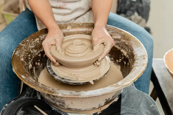 Обрезанный вид размытой женщины-ремесленницы в фартуке, делающей форму мокрой глины на вращающемся гончарном круге во время работы в керамической мастерской, квалифицированная концепция изготовления керамики — стоковое фото