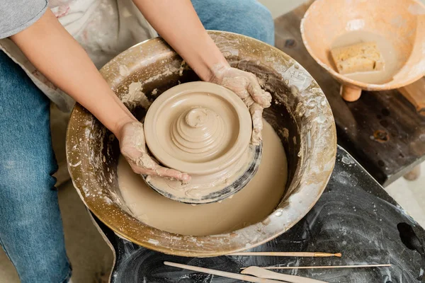 Vue du dessus de jeune artisan en tablier faisant forme d'argile humide sur la roue de poterie près du bol avec éponge et outils sur la table en atelier d'art, concept de fabrication de poterie habile — Photo de stock