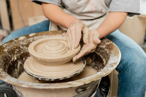 Обрезанный вид размытой женщины-ремесленницы в фартуке, делающей форму глины на вращающемся гончарном круге во время работы в классе керамики, квалифицированная концепция изготовления керамики — стоковое фото