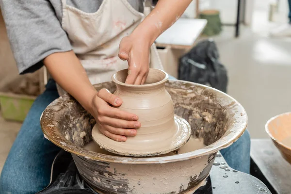 Vista cortada de oleiro fêmea jovem em vaso de argila de moldagem de avental e trabalhando com roda de cerâmica giratória em oficina de cerâmica turva no fundo, processo de criação de cerâmica — Fotografia de Stock