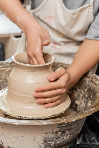 Обрезанный вид молодой женщины-ремесленницы в фартуке, делающей форму глиняной вазы и работающей с вращающимся керамическим колесом на столе в керамической мастерской, процесс создания керамики — стоковое фото