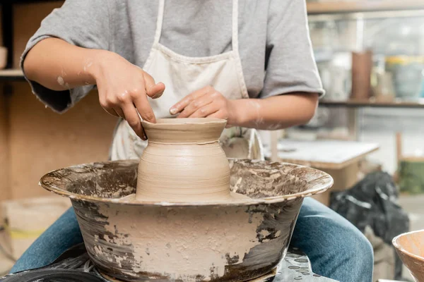 Vista recortada de ceramista jovem em avental e vestuário de trabalho fazendo forma de vaso de argila e trabalhando com roda de cerâmica em oficina de cerâmica turva, processo de criação de cerâmica — Fotografia de Stock