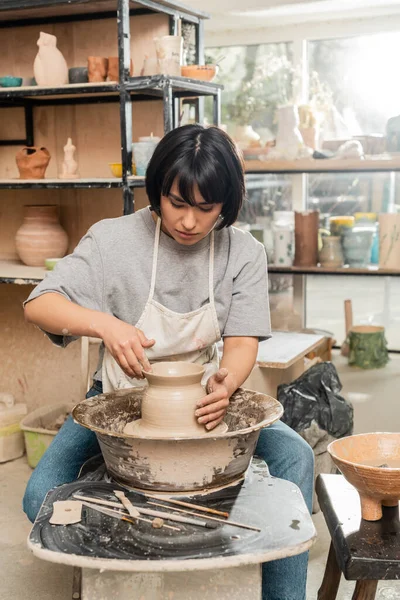Молодая азиатская брюнетка ремесленница в фартуке и рабочей одежде, делающая глиняную вазу и работающая на вращающемся гончарном круге в размытой керамической мастерской на заднем плане, процесс создания керамики — стоковое фото