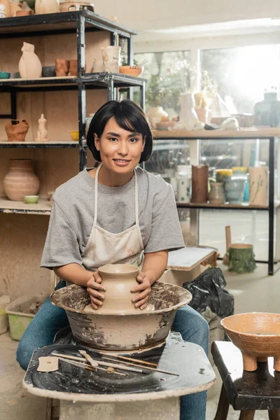 Улыбающаяся молодая азиатская ремесленница в фартуке делает форму глиняной вазы и смотрит в камеру, работая с гончарным кругом в размытой художественной мастерской, процесс создания керамики — стоковое фото