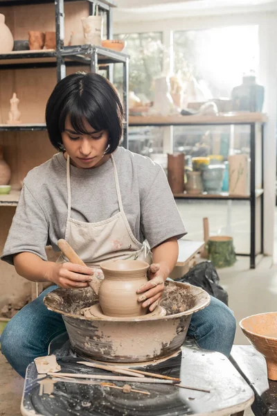 Young brunette asian female potter in apron holding wooden tool near clay while working with spinning pottery wheel in blurred ceramic workshop, pottery creation process — Stock Photo