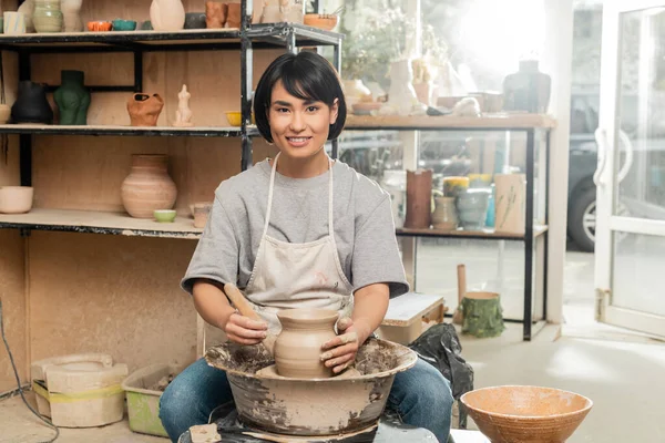 Souriant jeune brune asiatique femme artisan dans tablier regardant caméra tout en tenant outil en bois près de l'argile sur la roue de poterie en atelier de céramique à l'arrière-plan, façonnage de l'argile et processus de formage — Photo de stock