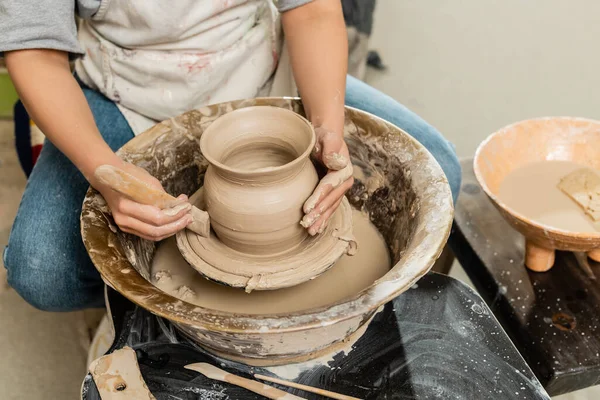 Cropped view of young female potter in apron making shape of clay vase with wooden tool on pottery wheel near bowl with water and sponge in art studio, clay shaping and forming process — Stock Photo
