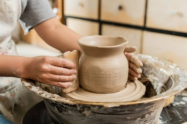 Обрізаний вид молодої жінки-художниці в фартусі, що робить глиняну вазу з дерев'яним скребком на керамічному колесі в розмитій керамічній майстерні, формування глини і процес формування — стокове фото