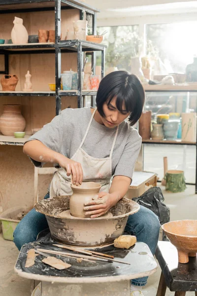 Junge brünette asiatische Handwerkerin in Schürze, die die Form einer Tonvase herstellt, während sie mit der Töpferscheibe in der Nähe von Schwamm und Holzwerkzeugen in der Keramikwerkstatt arbeitet, Formen und Formen von Ton — Stockfoto