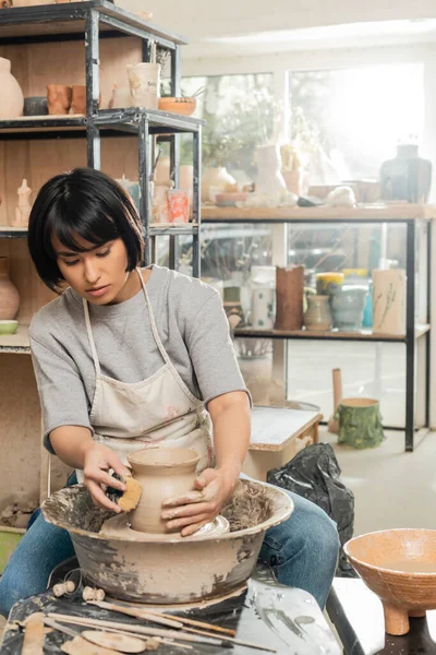 Young asian female artisan in apron making shape of clay vase with wet sponge and working with spinning pottery wheel in blurred ceramic workshop, clay shaping and forming process — Stock Photo