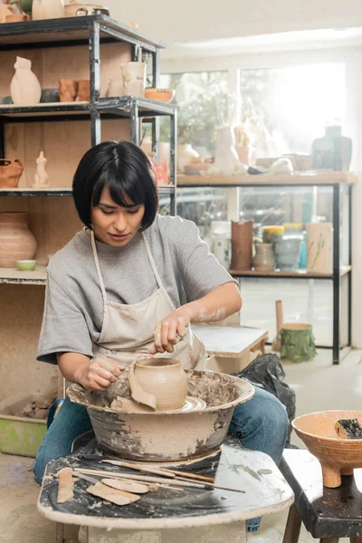 Brunette asian female artisan in apron cutting wet clay on spinning pottery wheel while working near wooden tools and bowl in blurred ceramic workshop, clay shaping and forming process — Stock Photo