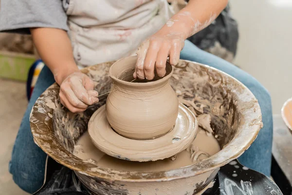 Vue recadrée de l'artisan brune dans un vase en argile de coupe de tablier sur une roue de poterie tout en travaillant dans un atelier de céramique à l'arrière-plan, façonnage et formage de l'argile — Photo de stock