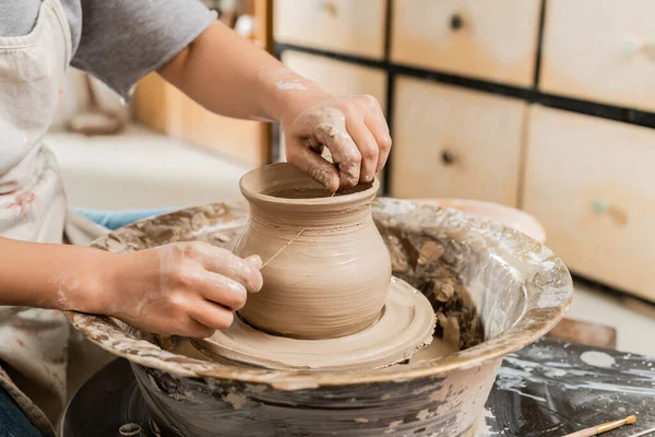 Обрізаний вид молодої жінки-художниці в фартусі, що розрізає мокру глиняну вазу на обертовому керамічному колесі на столі в розмитій керамічній майстерні, формування глини і процес формування — стокове фото