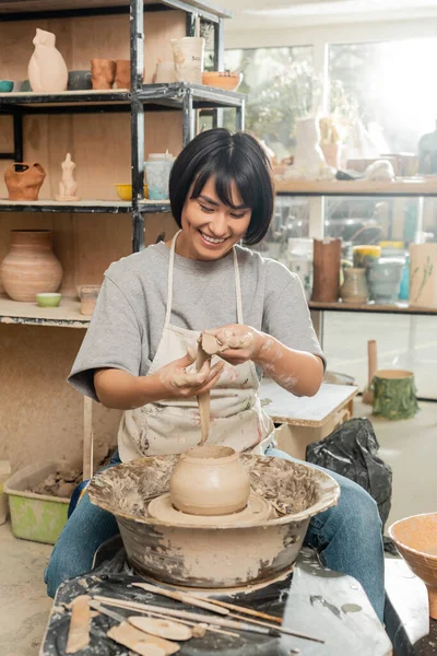 Positivo giovane asiatica vasaio femminile in grembiule tenendo argilla vicino vaso sulla ruota di filatura ceramica e strumenti in legno e ciotola in studio di ceramica sfocata, produzione e processo di ceramica artigianale — Foto stock