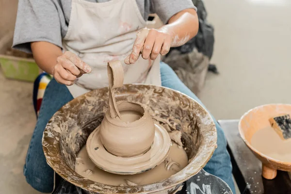 Vista ritagliata di artigiano fmale in grembiule che tiene argilla mentre fa vaso sulla ruota della ceramica di filatura vicino alla ciotola offuscata con acqua in laboratorio di ceramica, produzione e processo di ceramica artigianale — Foto stock