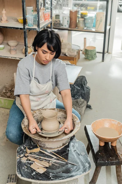 Jovem asiática ceramicista feminina em avental moldando vaso de barro com ferramentas na roda de cerâmica giratória perto de esponja e tigela com água no estúdio de cerâmica, produção e processo de cerâmica artesanal — Fotografia de Stock