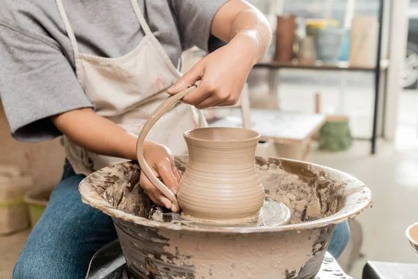 Vista cortada de oleiro jovem fêmea em avental fazendo jarro de barro enquanto trabalhava com roda de cerâmica em oficina de cerâmica borrada no fundo, produção e processo de cerâmica artesanal — Fotografia de Stock